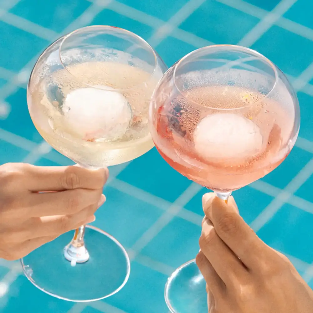 Two hands clinking glasses of Veuve Clicquot Rich on Ice and Rich on Ice Rosé with ice balls, set against a bright blue tiled pool background.
