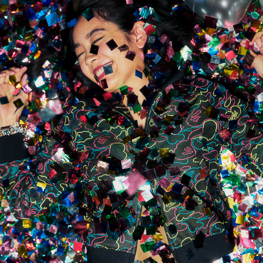 Person joyfully lying in colorful confetti, wearing a patterned jacket.