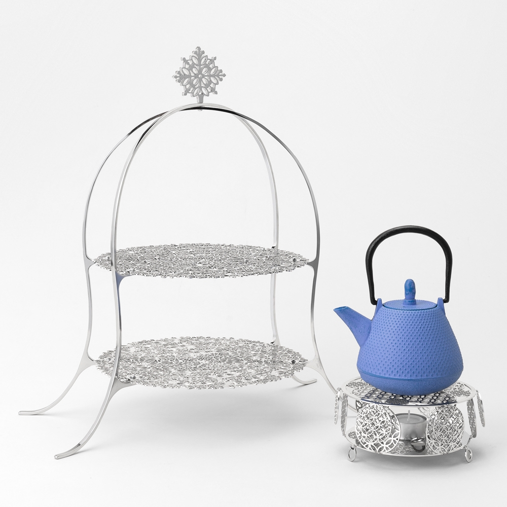 Ornate two-tier silver stand with intricate designs, next to a blue teapot on a matching silver base.