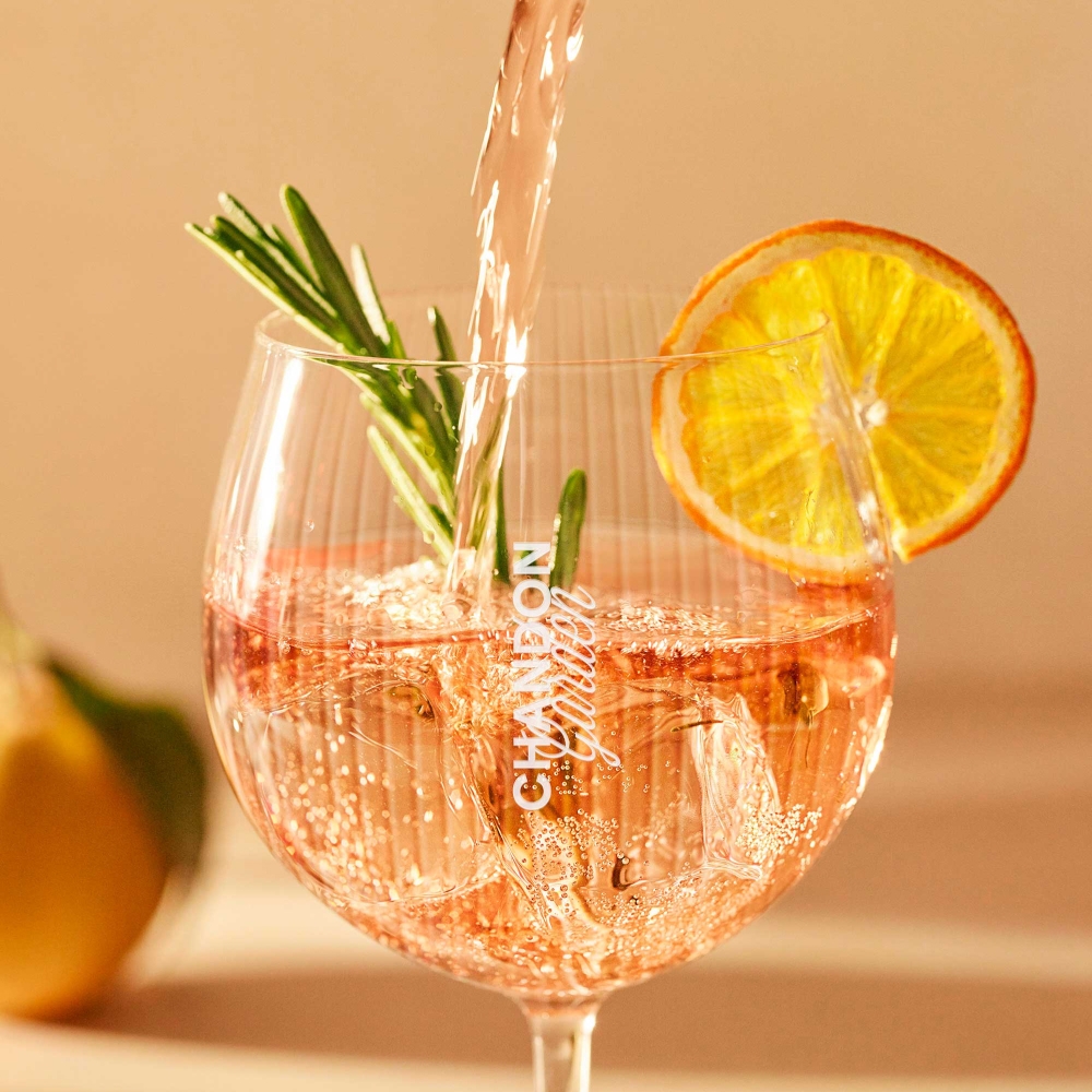 A glass of sparkling wine, CHANDON  Garden Spritz with ice cubes rosemary sprig and orange slice garnish, being poured, set against a warm, blurred background.