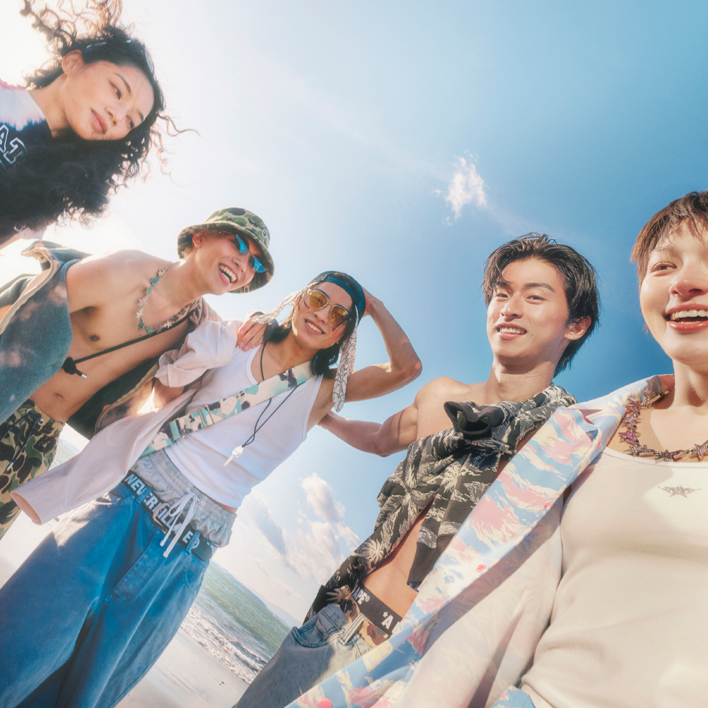 A group of five friends smiling and posing on a sunny beach, wearing casual beachwear, with a clear blue sky in the background.