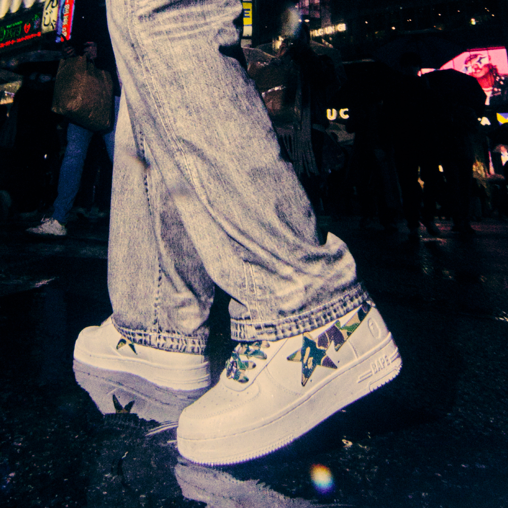 Person wearing white sneakers with a colorful design stands on a wet street at night, with reflections visible on the ground.