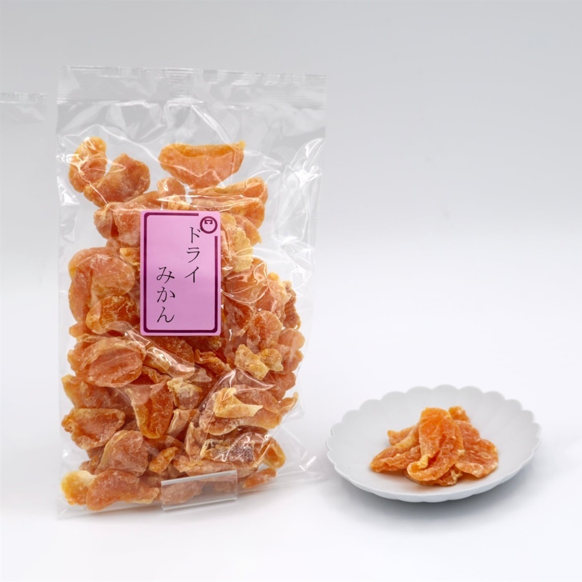 A bag of dried tangerine slices with pink Japanese labeling, next to a white plate containing a few slices, on a white background.