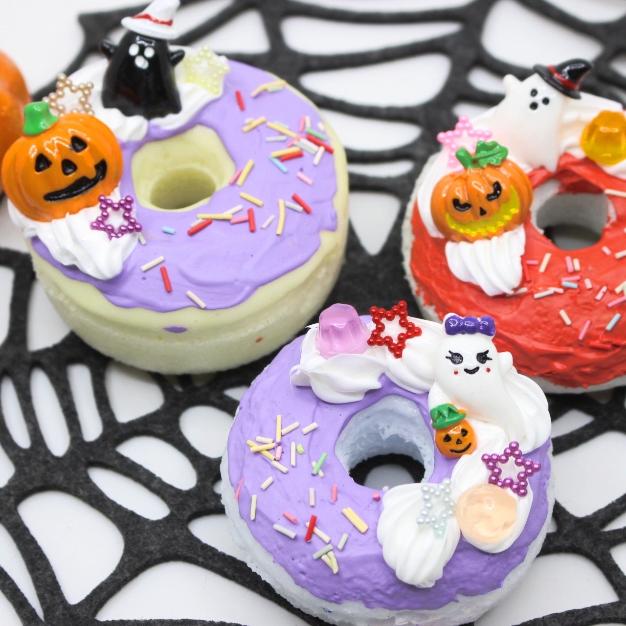 Three Halloween-themed donuts with colorful icing, sprinkles, and decorations like pumpkins, ghosts, and stars, on a spider web backdrop.