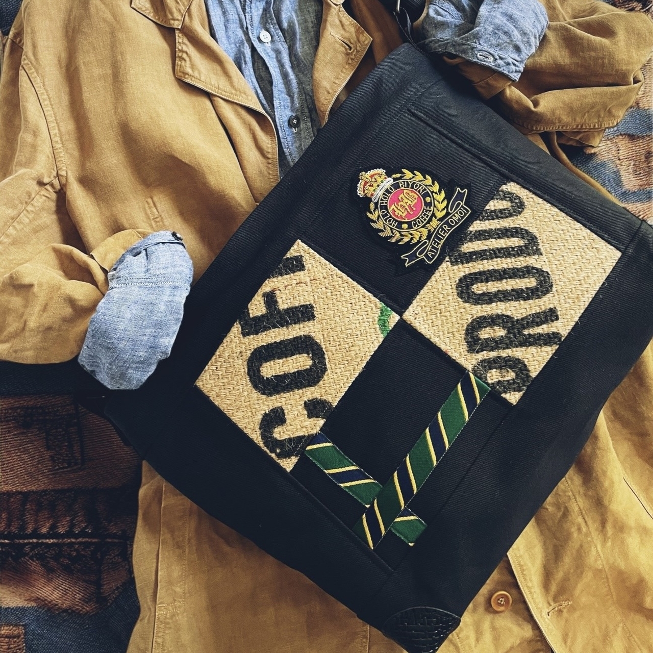 A black bag with coffee-themed patches and a striped green tie, placed on a brown jacket and denim shirt.