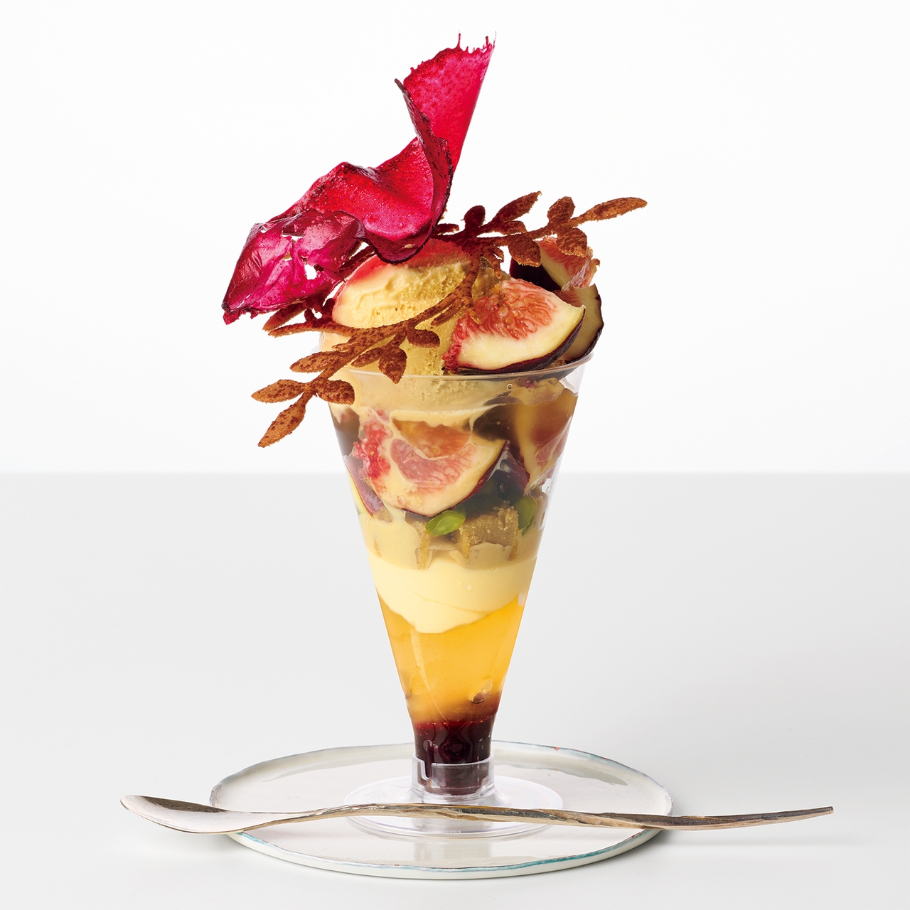 A layered dessert parfait in a glass with figs, cream, a red decorative topper, and a spoon beside it on a clear plate.