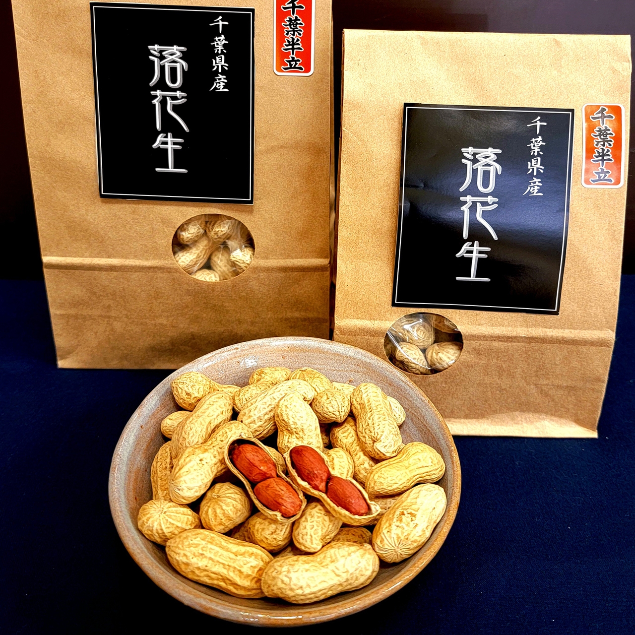A bowl of peanuts in shells, some cracked open to reveal red skins, in front of two brown bags with black labels featuring Japanese text.