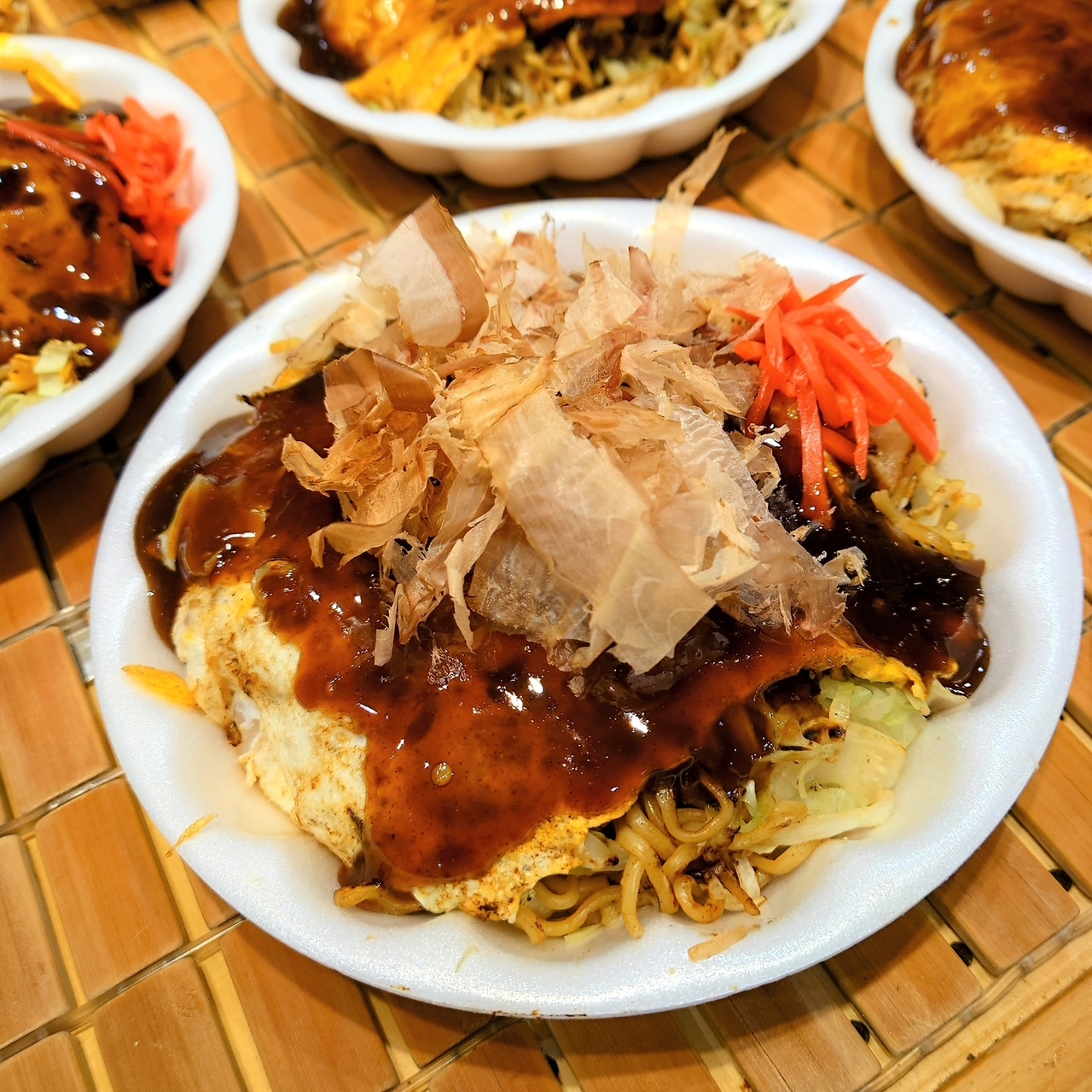 A dish of okonomiyaki topped with bonito flakes, sauce, and pickled ginger, served on a white plate placed on a bamboo mat.