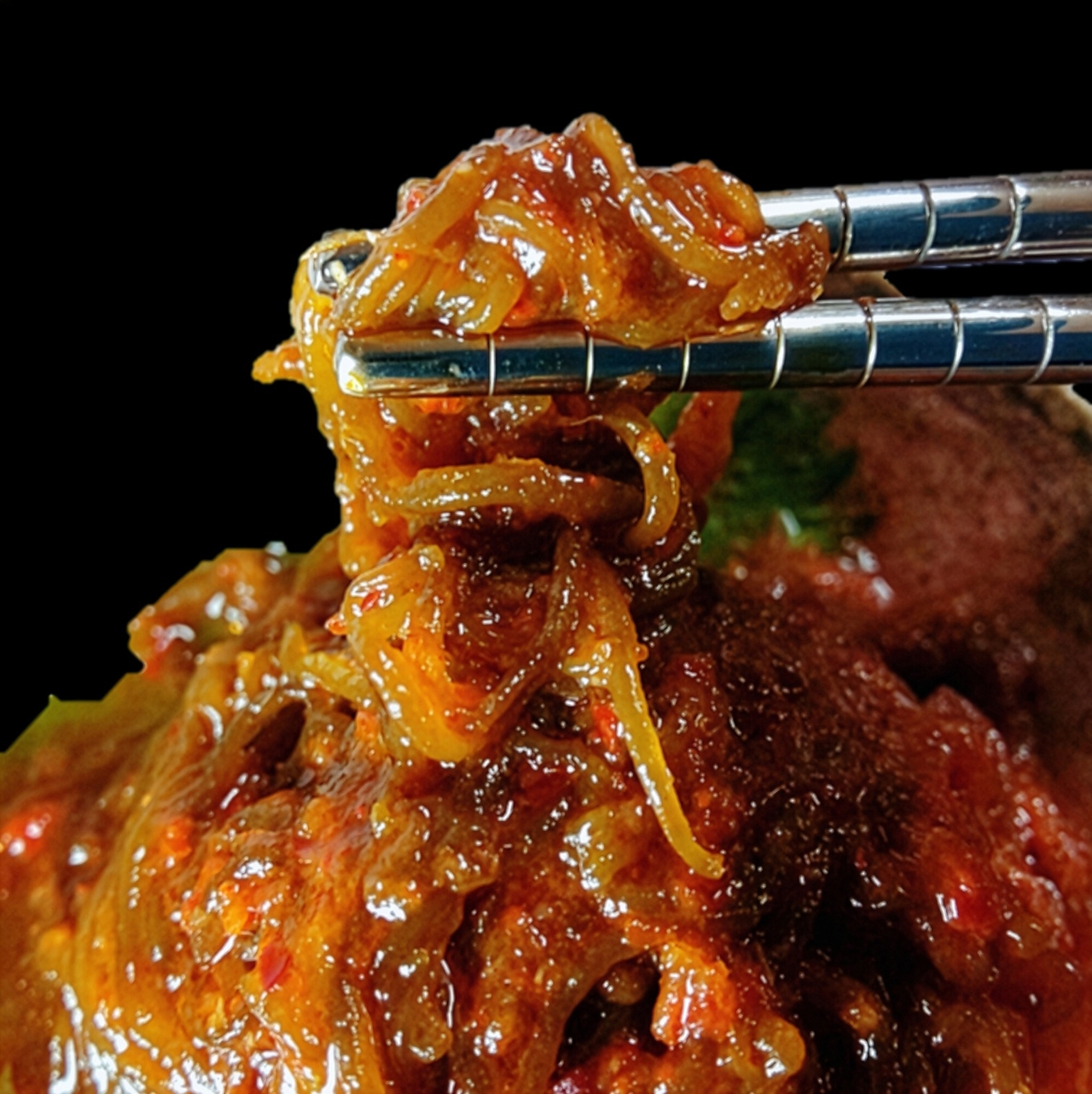 Chopsticks holding a portion of spicy, saucy shredded meat dish with a glistening texture against a black background.