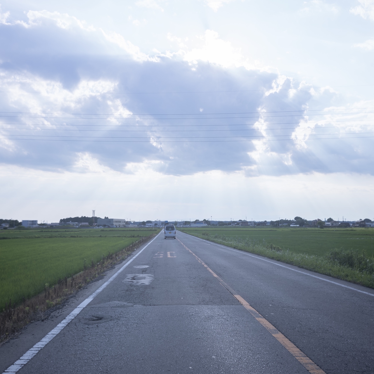 A rural road stretches into the distance under a sky with sun rays streaming through clouds, bordered by green fields on both sides.