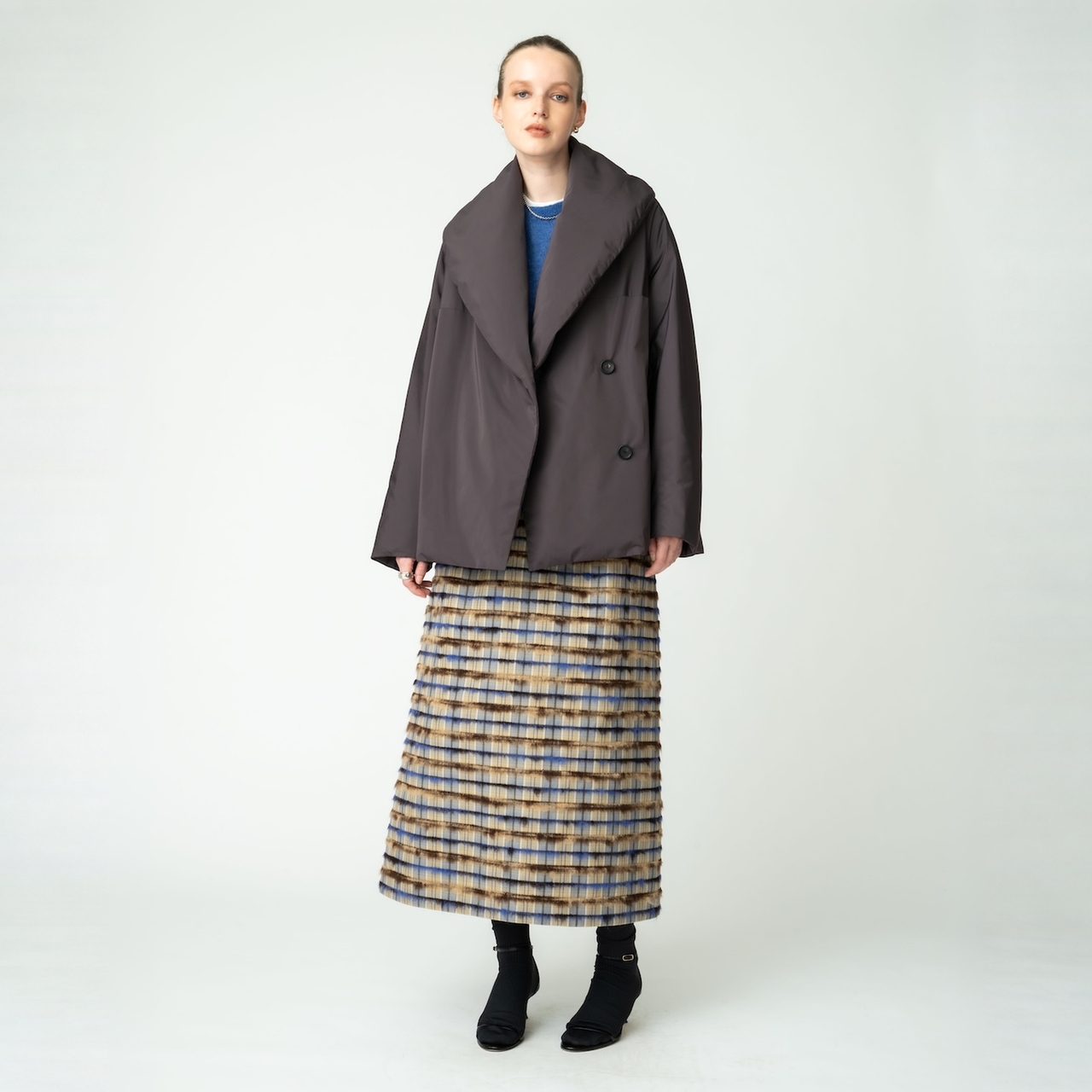 無地の背景の前に立つ、オーバーサイズのパープルのコート、ブルーのトップス、ロングチェック skirts（チェック柄ロングスカート）を着た女性。
