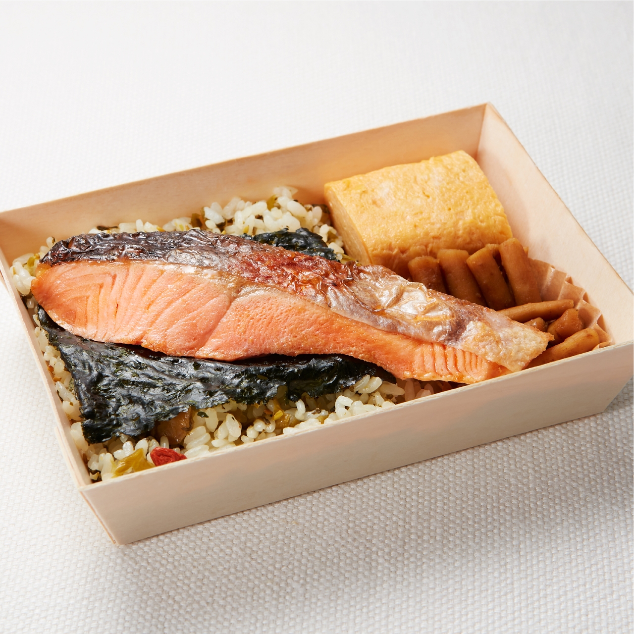 A bento box with grilled salmon on rice, garnished with seaweed, accompanied by tamagoyaki and lightly pickled vegetables, placed on a light surface.