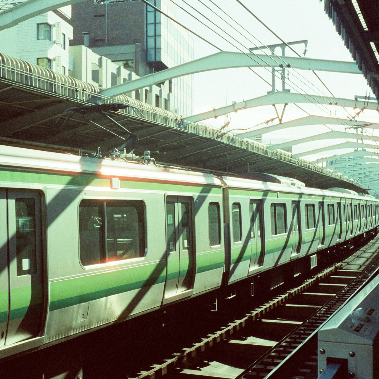 A green-striped train at a city station under a bridge, with sunlight casting shadows on the platform and tracks.