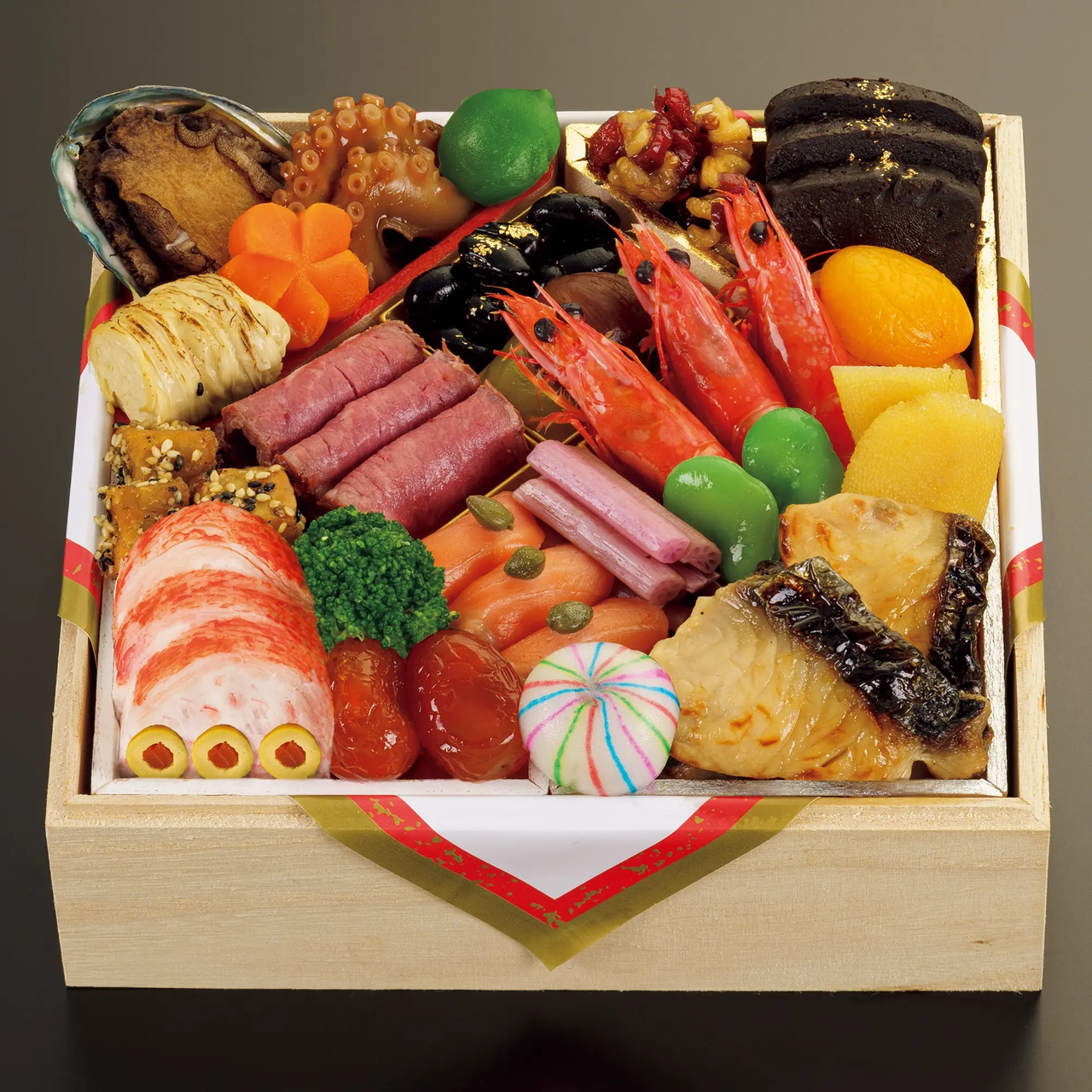 A colorful Japanese osechi bento box with shrimp, grilled fish, rolled meats, vegetables, beans, and mochi, arranged in a square wooden container.