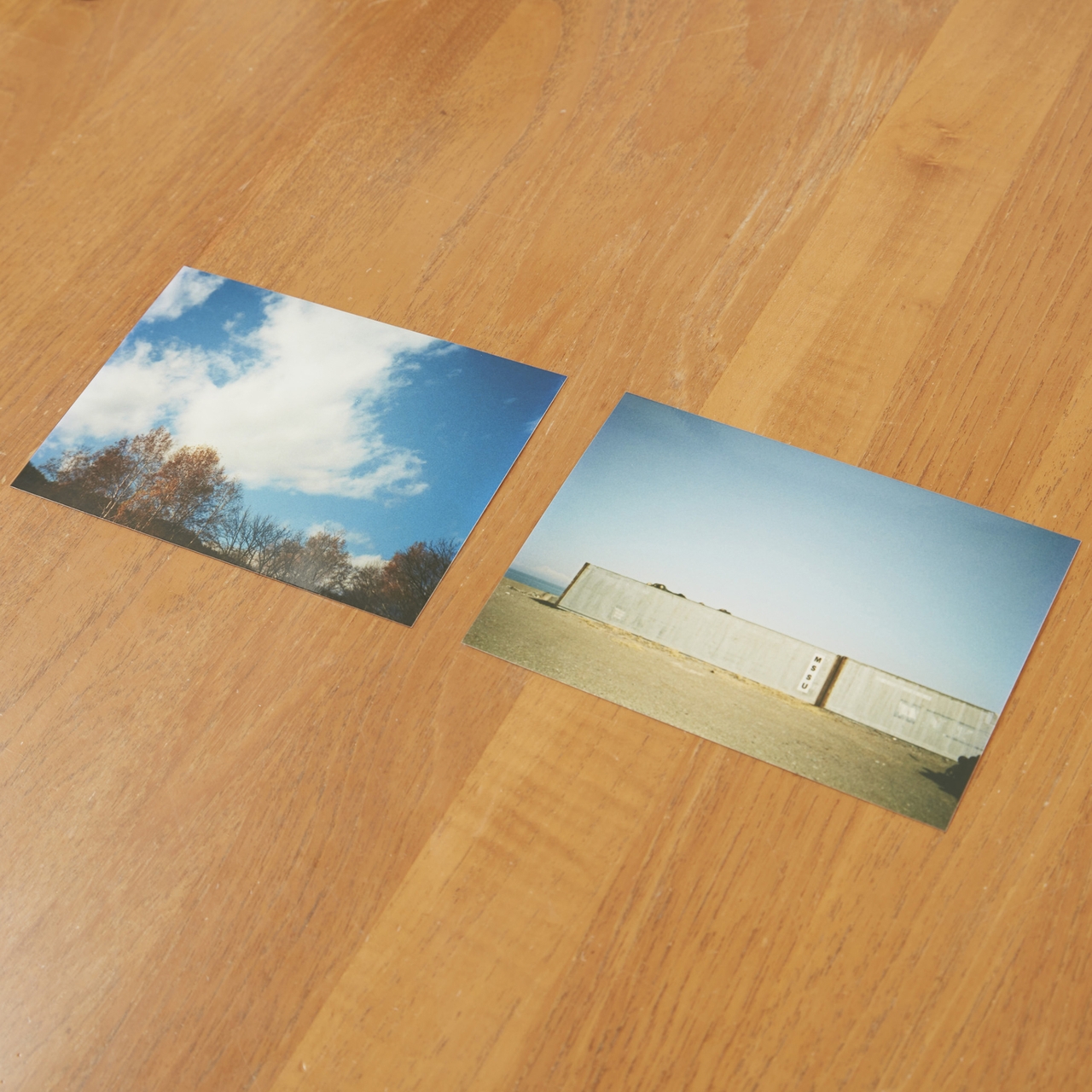 Two photographs on a wooden surface: one of a cloudy sky with trees, the other of a large, flat building under a clear blue sky.