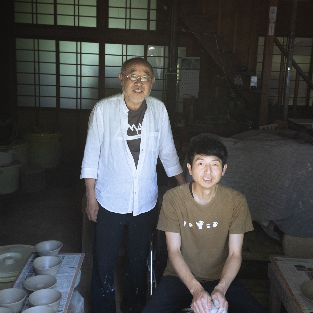 Two men in a pottery workshop, one standing and one seated, surrounded by pottery items. Traditional Japanese interior with sliding doors.