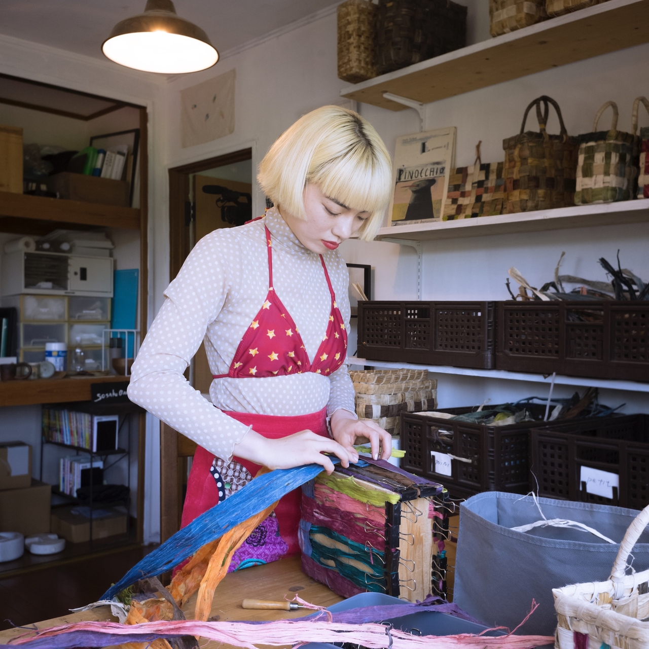 A person with blonde hair works on a colorful craft project in a cozy, organized room filled with baskets and materials.