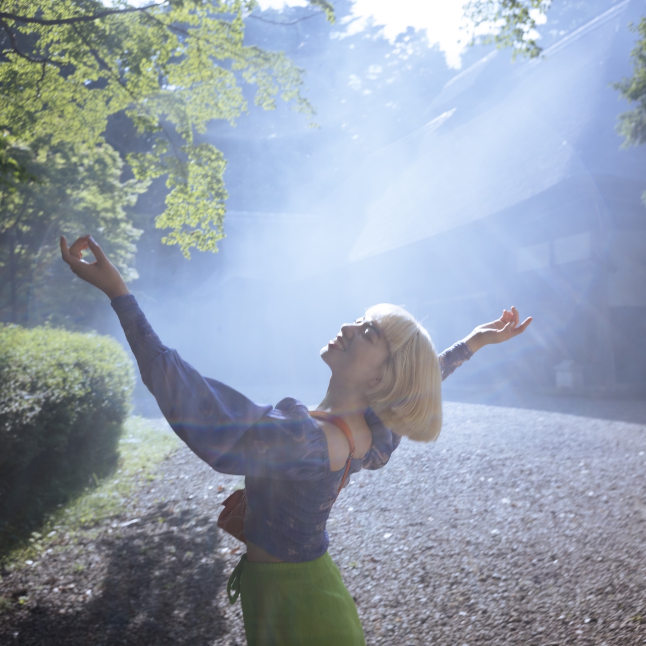 Person with short blonde hair joyfully raising arms in a sunlit, misty forest clearing.