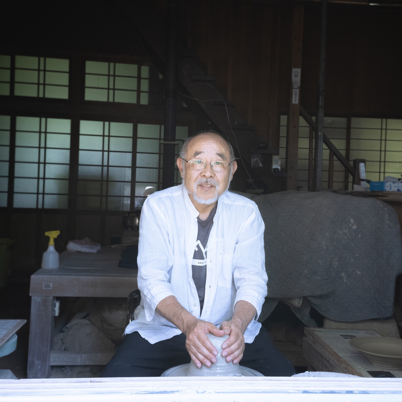 Elderly man in a white shirt sitting indoors, shaping clay on a pottery wheel, with traditional wooden doors in the background.