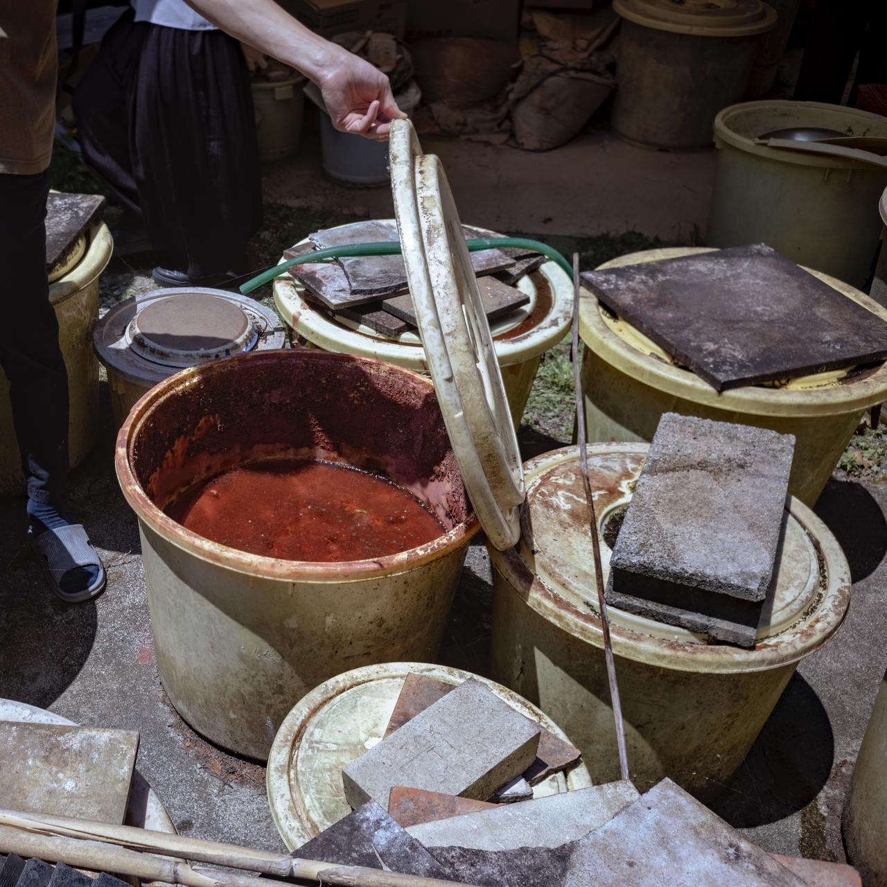 Person lifting the lid of a large container filled with red liquid, surrounded by other covered containers and stone slabs outdoors.