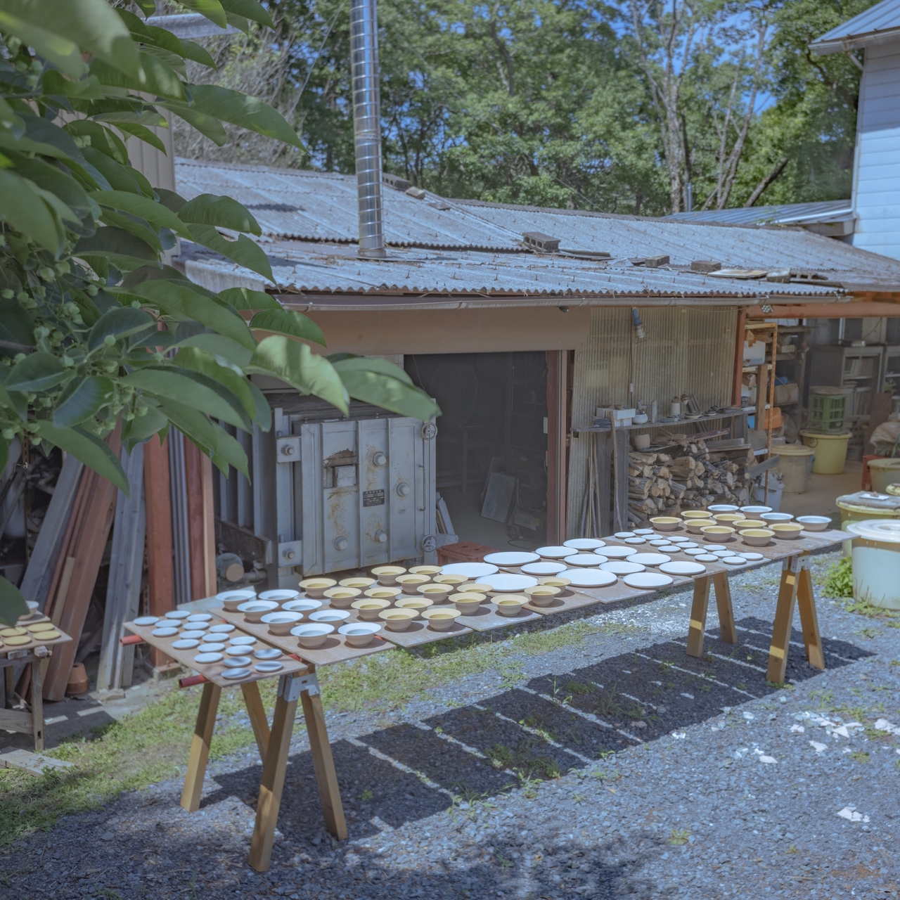 Wooden tables outside a rustic shed display various ceramic plates and bowls under a clear blue sky, surrounded by greenery.
