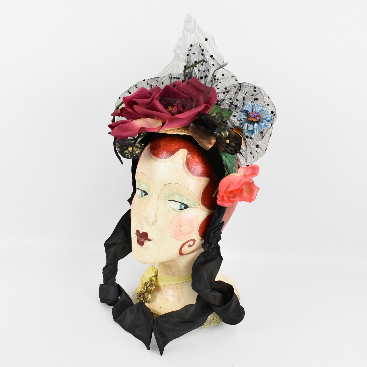 Vintage mannequin head with red hair, wearing a decorative hat adorned with large roses, lace, and ribbons.