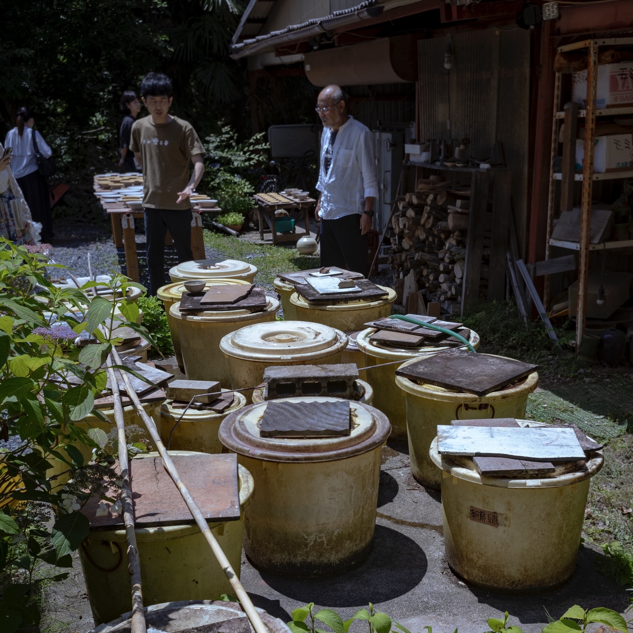 People standing near large yellow ceramic pots with lids, outside a rustic building surrounded by greenery.