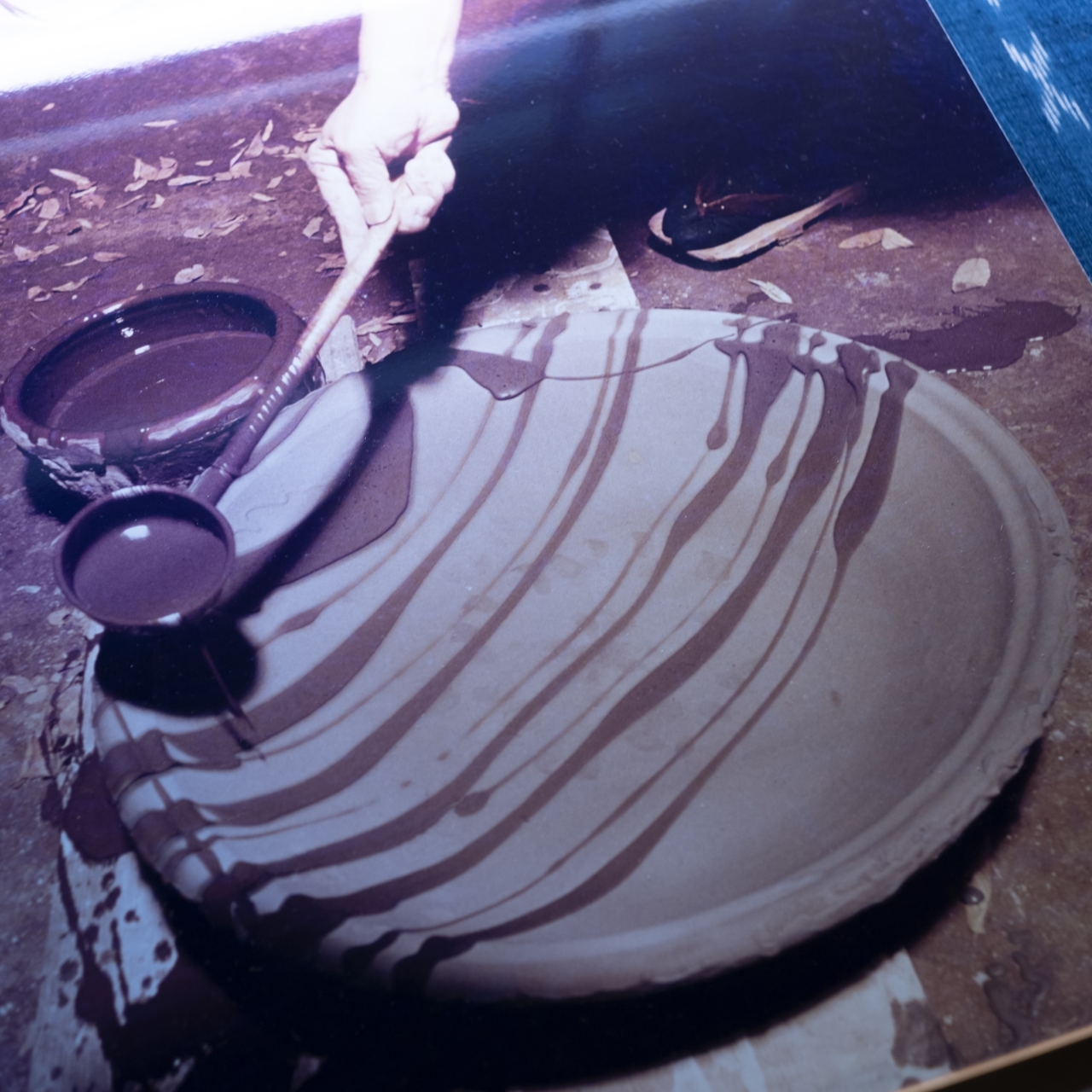 Hand pouring liquid clay onto a large, flat, round ceramic piece, creating striped patterns. Two bowls of clay are beside the piece.