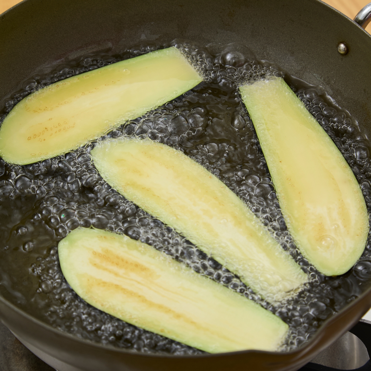 Slices of eggplant frying in hot oil in a black pan, with bubbles forming around the edges.