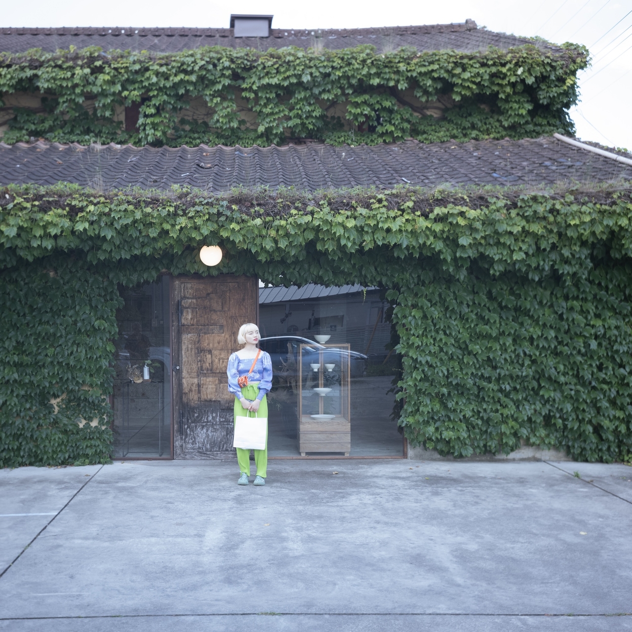 Person standing in front of a building covered in lush green ivy, holding a bouquet. The building has a rustic wooden door and tiled roof.