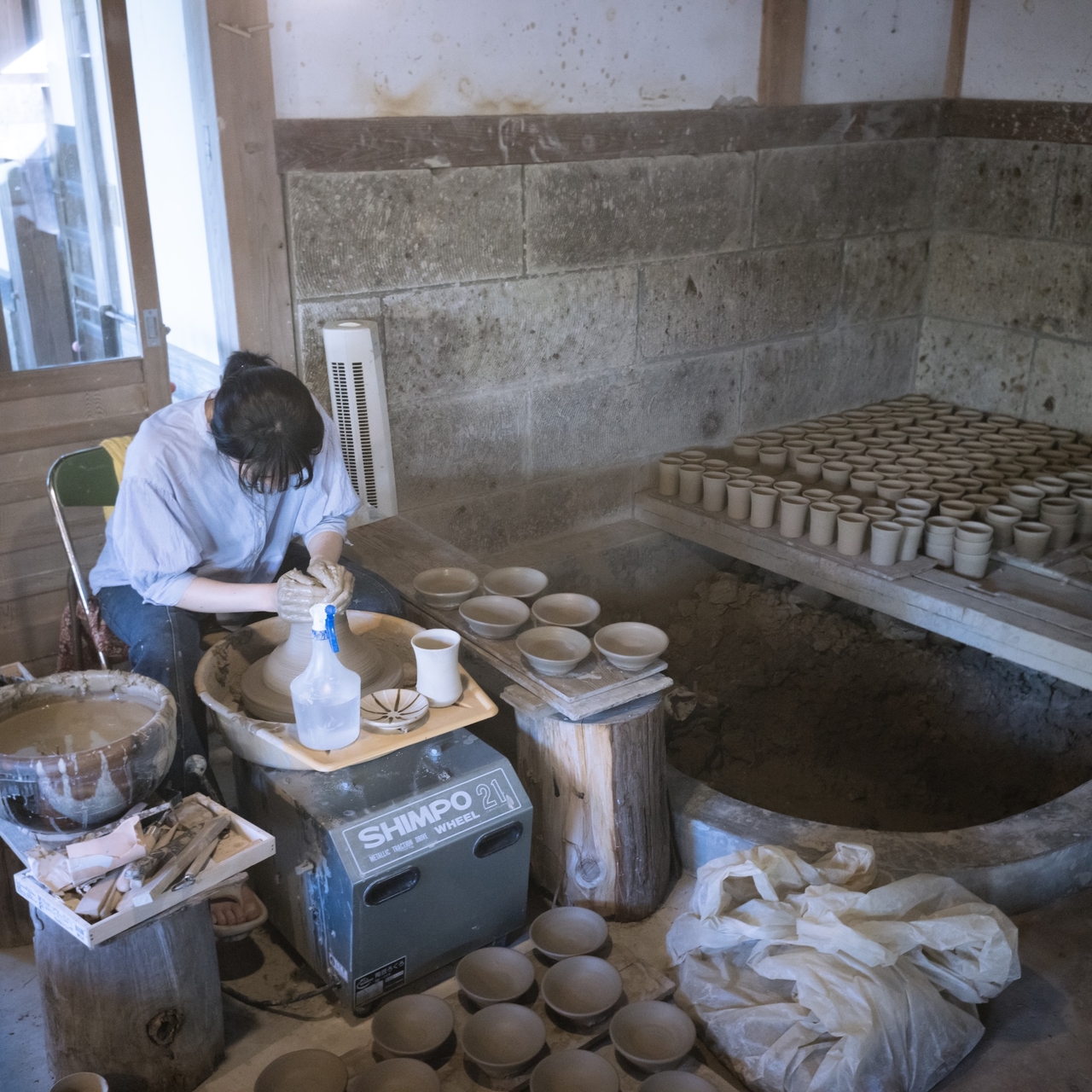 A person is crafting pottery on a wheel in a workshop, surrounded by clay pots and tools, with shelves filled with finished pieces.