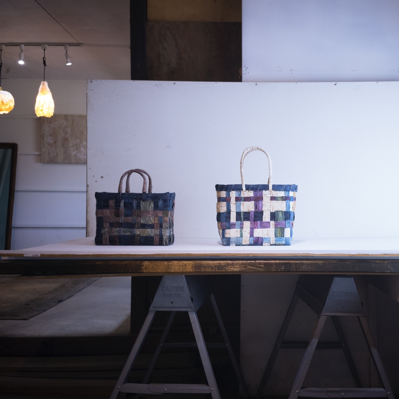 Two woven baskets with handles, featuring a checkered pattern, displayed on a wooden table in a softly lit room.
