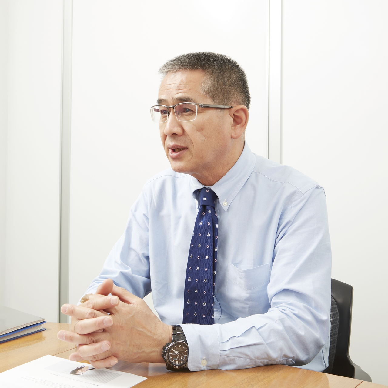 Man in glasses and blue shirt with patterned tie sits at a desk, speaking, with hands clasped. White background with a document in front.