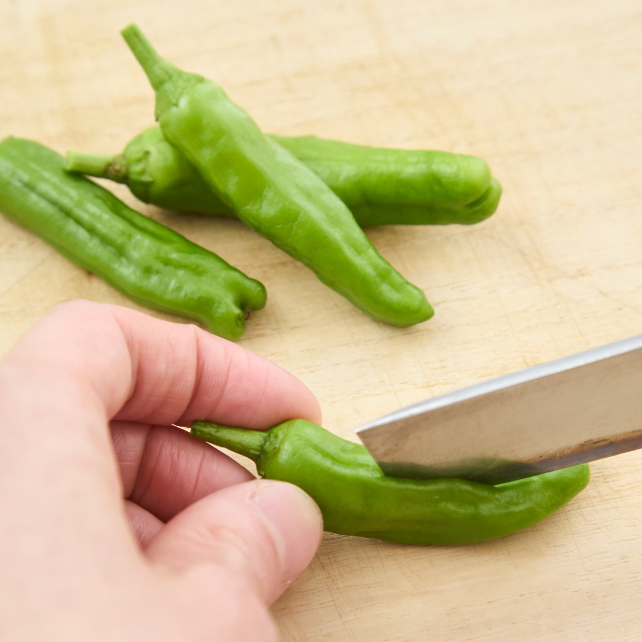 A hand slicing a fresh green chili pepper with a knife on a wooden cutting board, with three more peppers in the background.