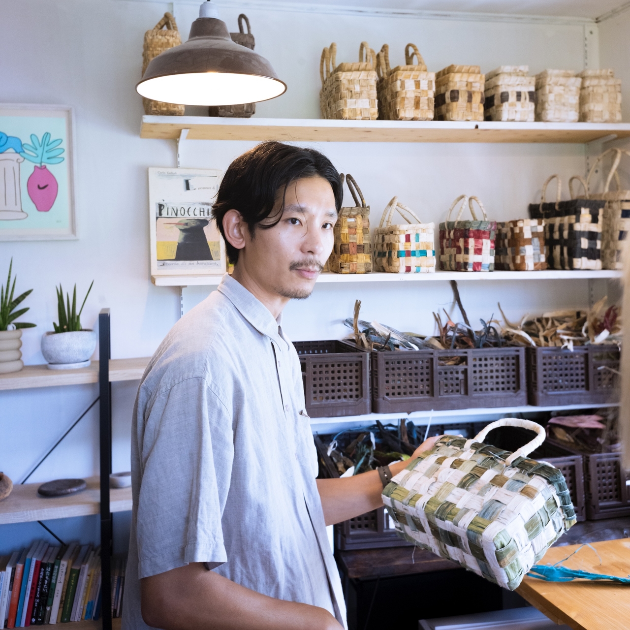 A person in a light shirt holds a woven basket in a room filled with various baskets and plants, under a hanging lamp.