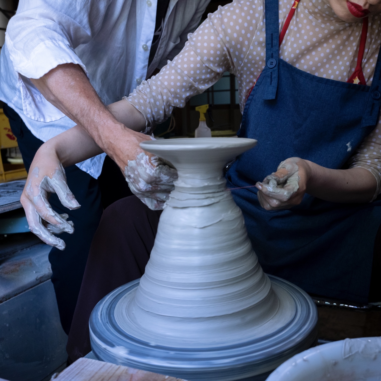 Two people shaping clay on a pottery wheel, hands covered in clay. One person wears a navy apron, the other a white shirt. They are trying to cut the final clay pottery