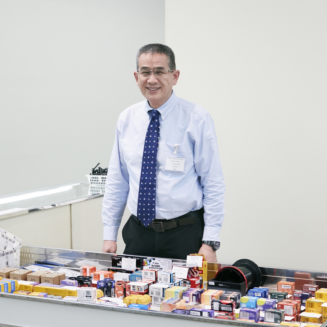 A man in a shirt and tie stands behind a counter with various boxed products in a bright, clean setting.
