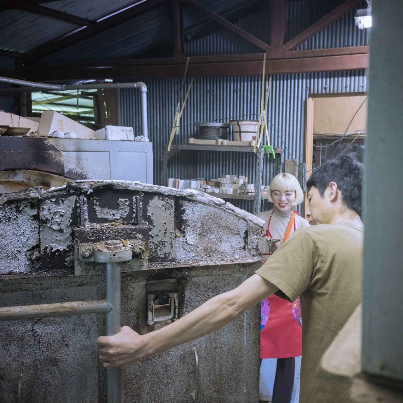 Two people in a workshop, one operating a large kiln. The setting is industrial with shelves and metal walls in the background.