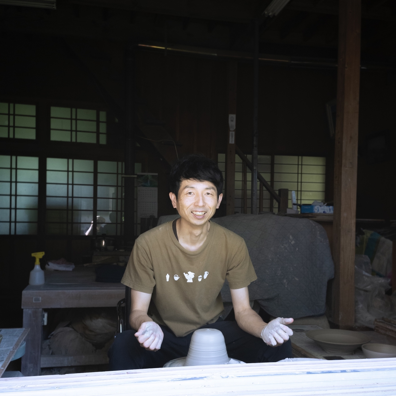 A person smiling while sitting at a pottery wheel in a dimly lit workshop, wearing a brown T-shirt and gloves. The man trying to start pottery.