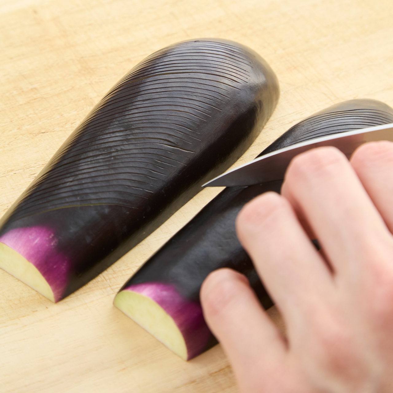 A hand slicing a shiny, dark purple eggplant with a knife on a wooden cutting board.