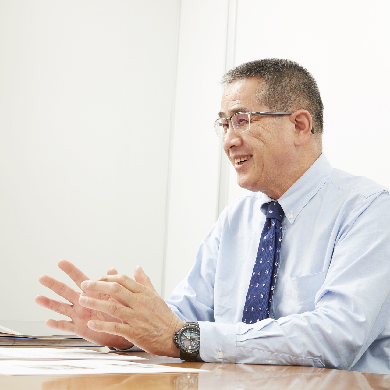 Smiling man in glasses and a blue shirt with a tie, gesturing with his hands while sitting at a table in a bright room.