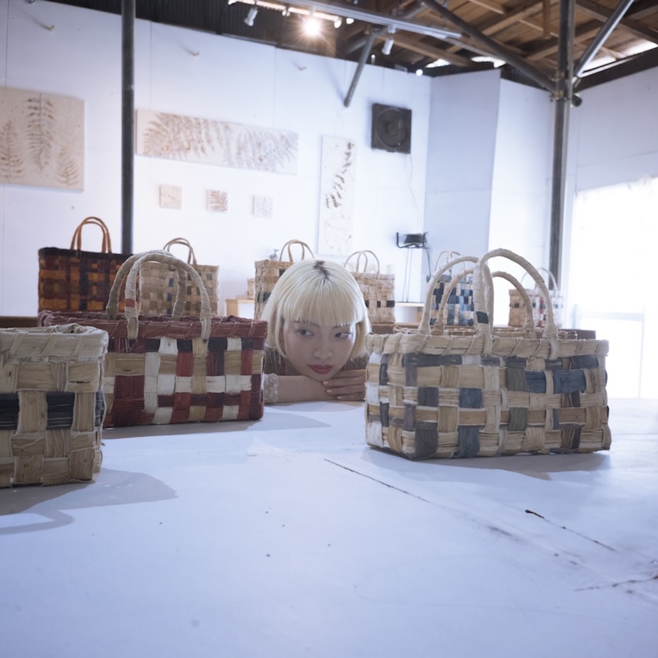 Person with blonde hair peeking over a table displaying woven baskets in an art studio.
