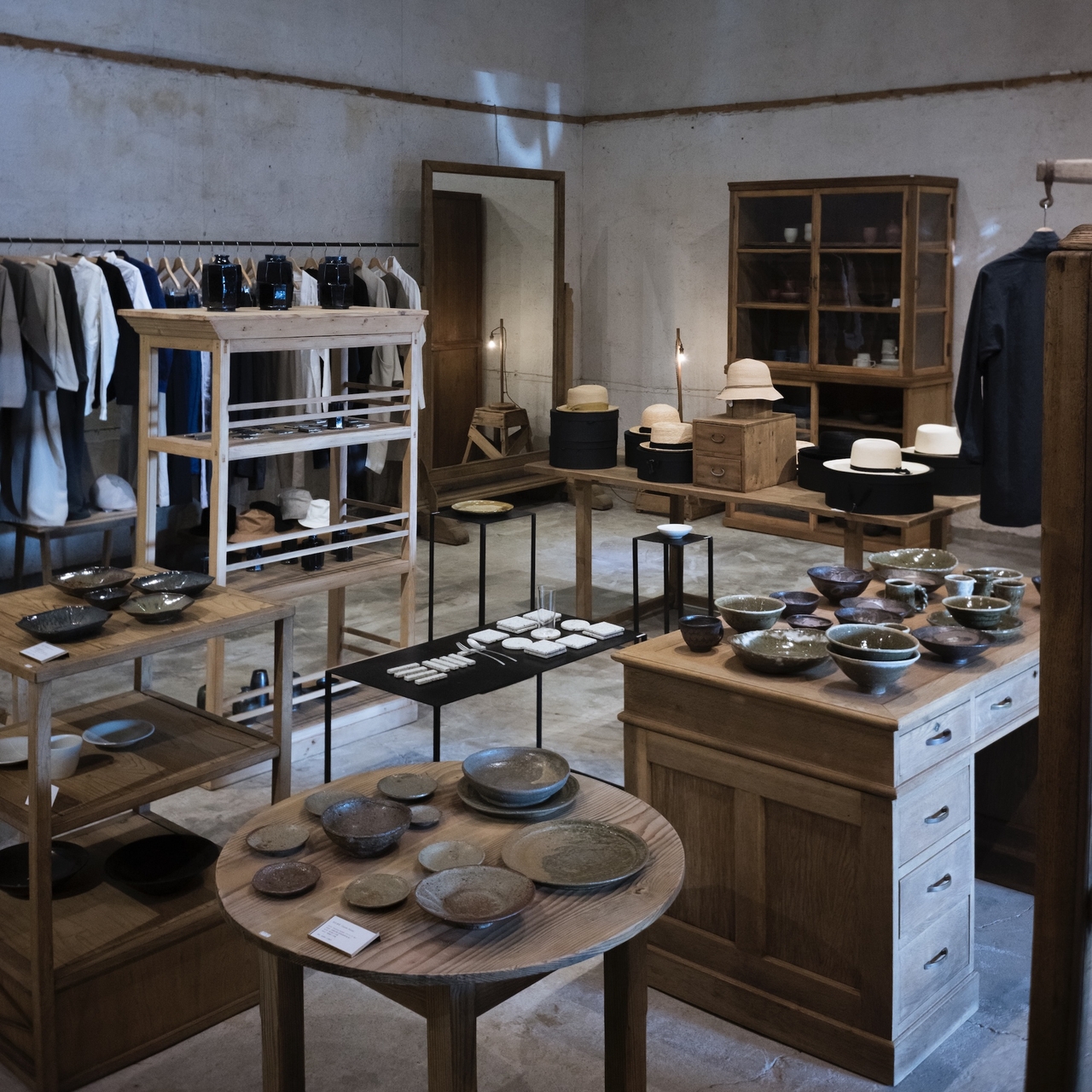 A cozy boutique interior with wooden shelves displaying pottery, clothing, and hats. Earthy tones and soft lighting create a warm atmosphere.