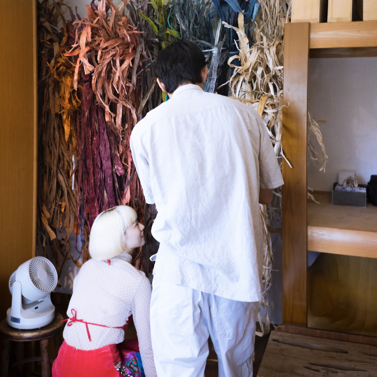 Two people in a room with colorful dried plants on the wall. One is crouching, and the other is standing, both wearing white clothing.