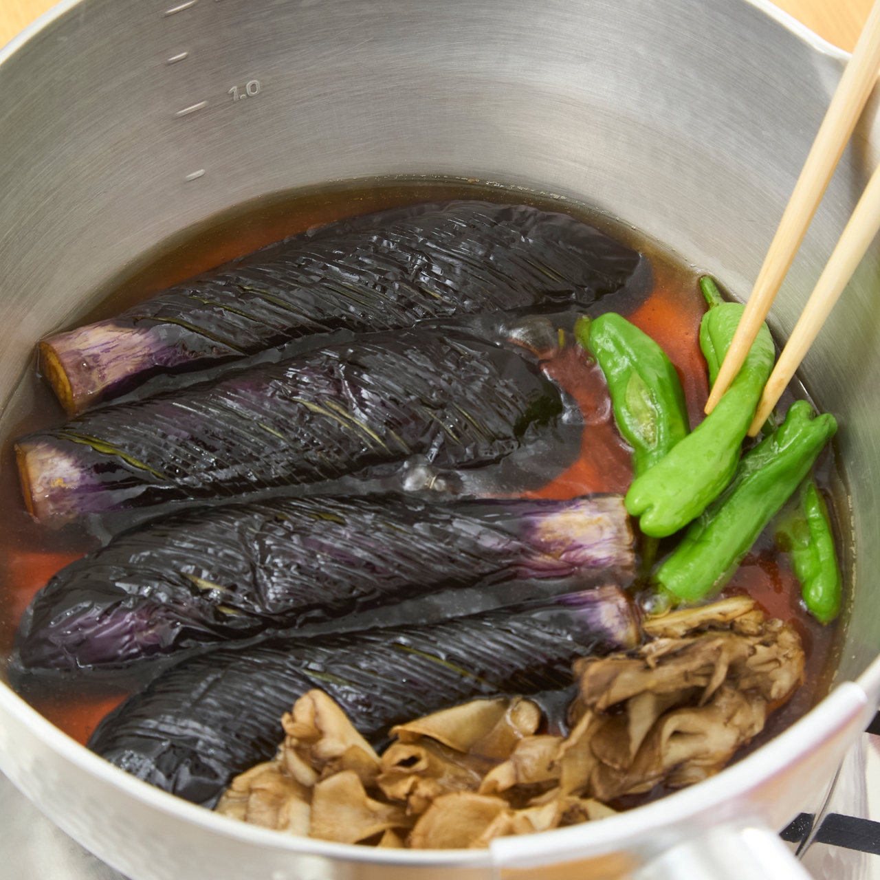 Eggplants simmering in a pot with a red sauce, green peppers, and mushrooms, being stirred with chopsticks.