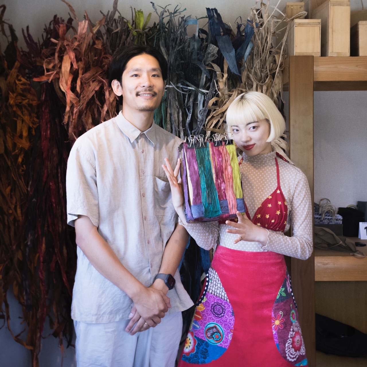 Two people stand in a colorful workshop, holding dyed fabric samples. One wears a vibrant apron, and dried plants hang in the background.