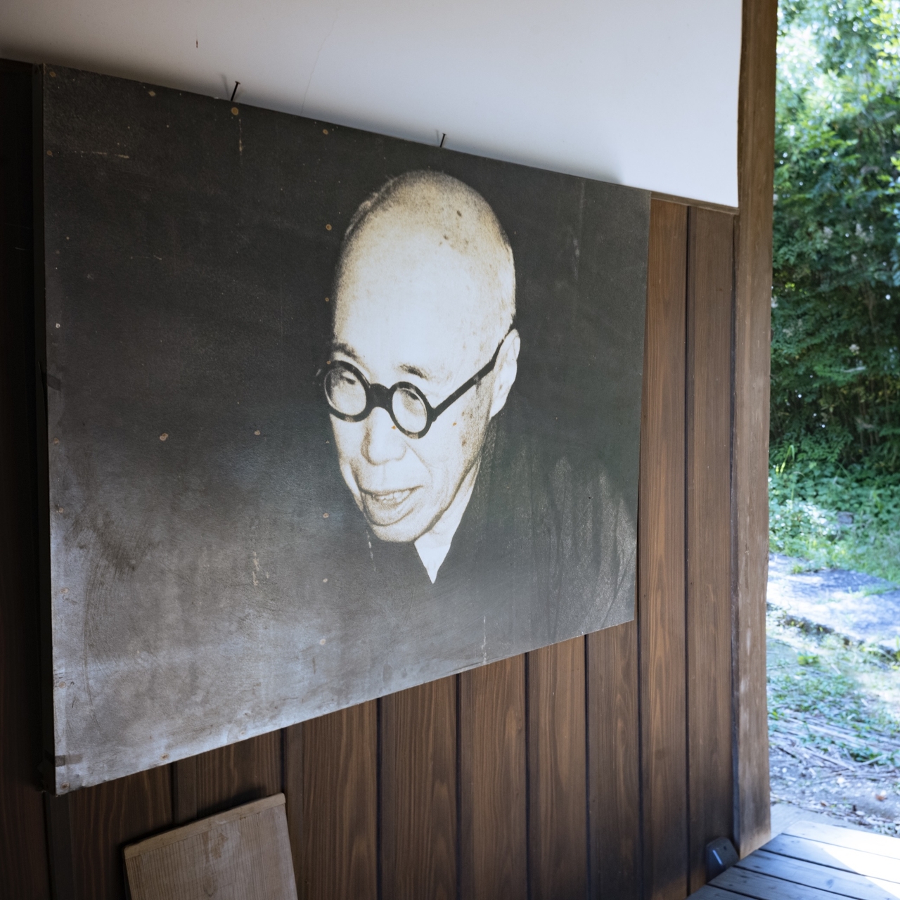 Black and white portrait of a man with glasses on a wooden wall, next to an open doorway with greenery outside. Sennorikyu's photo is hung on the wall.