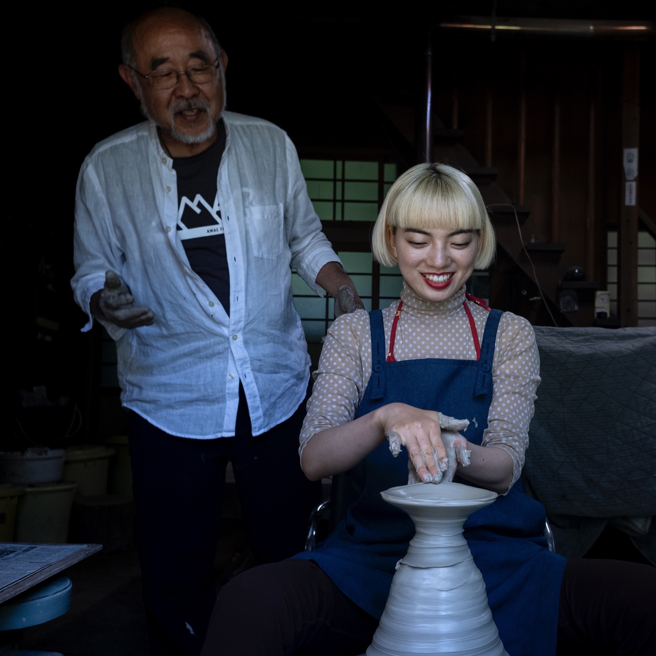 A woman in an apron shapes clay on a pottery wheel, smiling, while an older man stands nearby, guiding her in a dimly lit room.