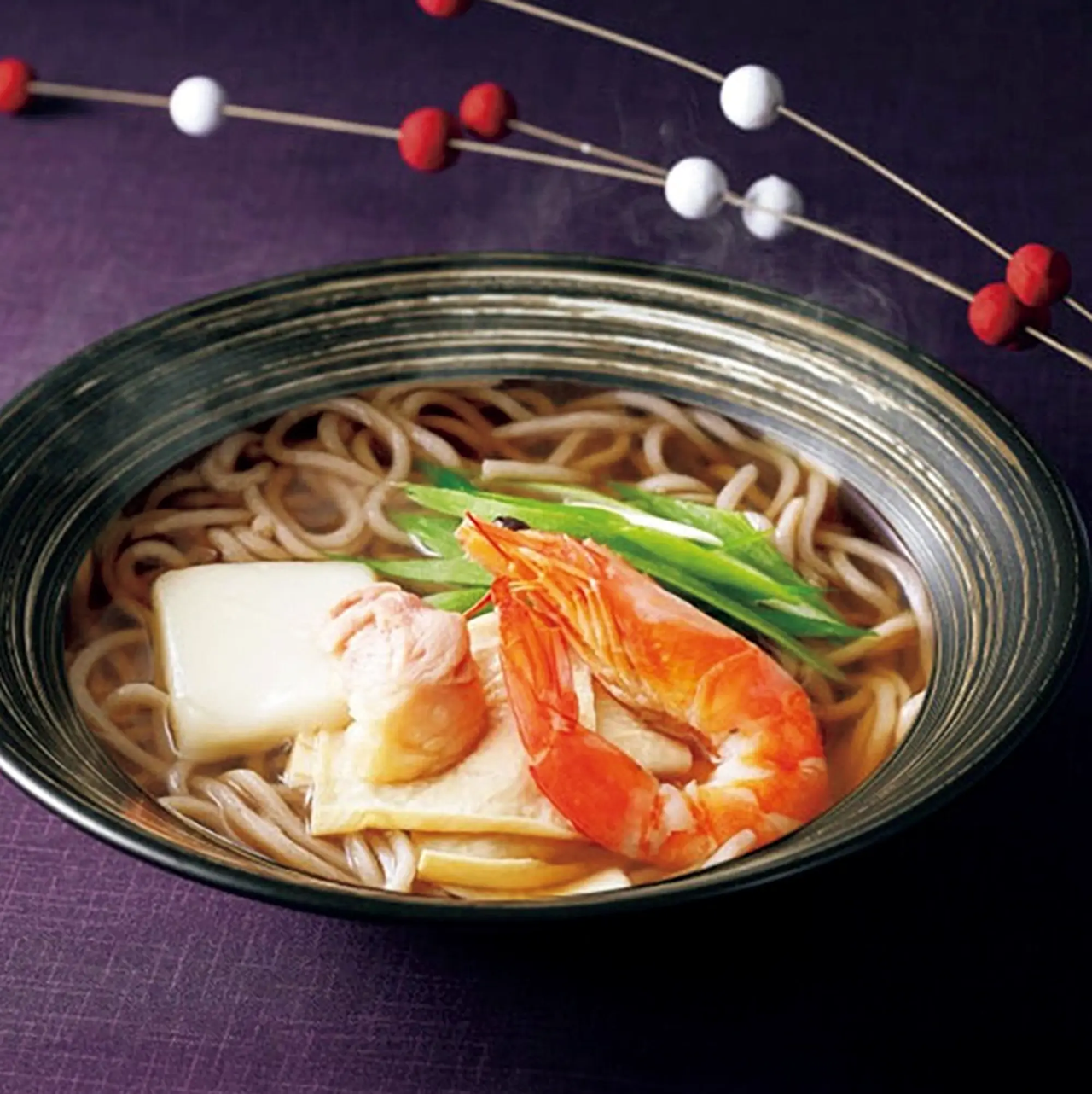 Bowl of soba noodles with shrimp, tofu, and green onions in broth, garnished with decorative red and white beads in the background.