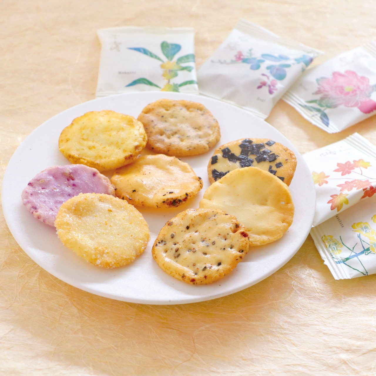 A white plate with assorted colorful rice crackers next to decorative floral packaging on a light textured background.